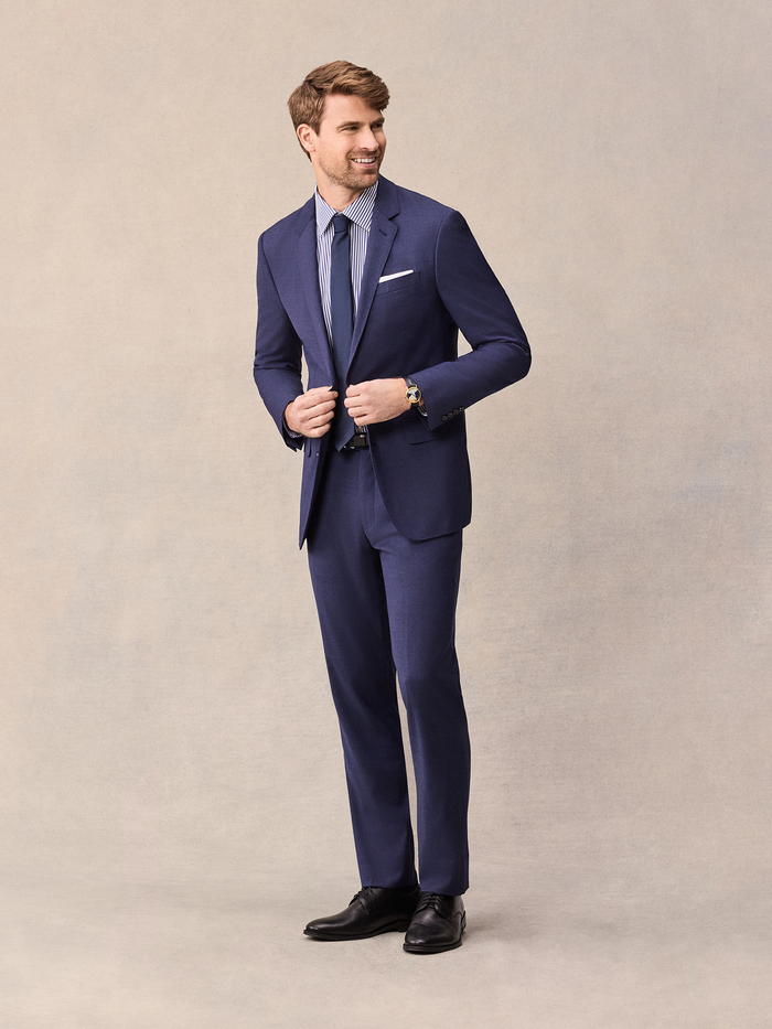 Man in a blue suit buttoning the jacket, three-quarter view on a beige backdrop
