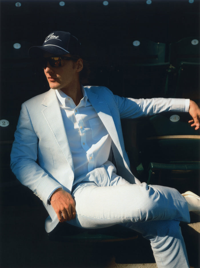 Man in a blue and white seersucker suit seated in baseball stadium stands, wearing a cap and sunglasses.