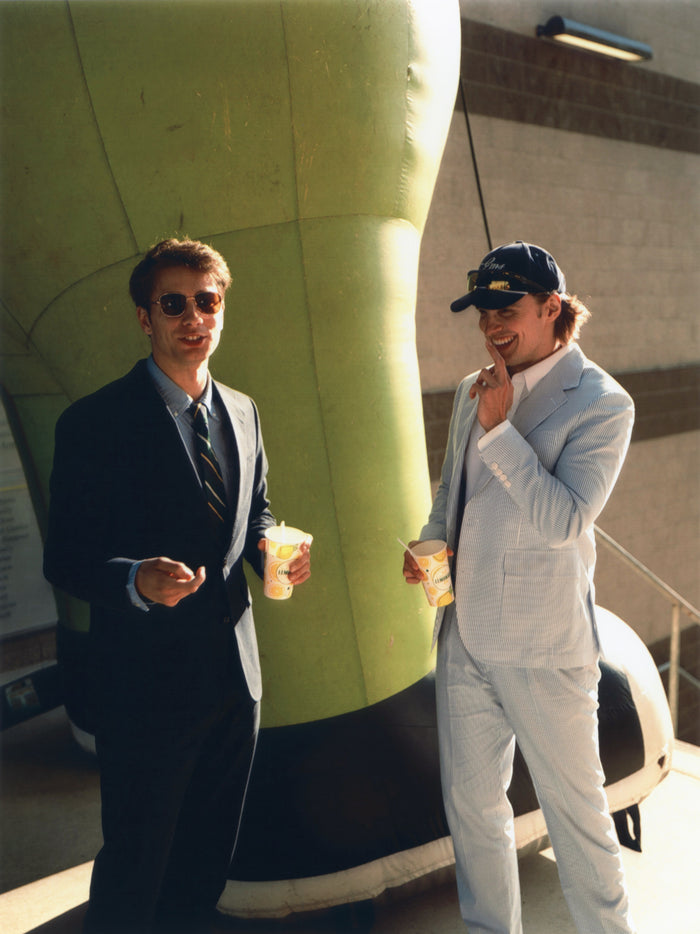 Two men holding drinks by a green inflatable, one in a blue and white seersucker suit, one in navy.