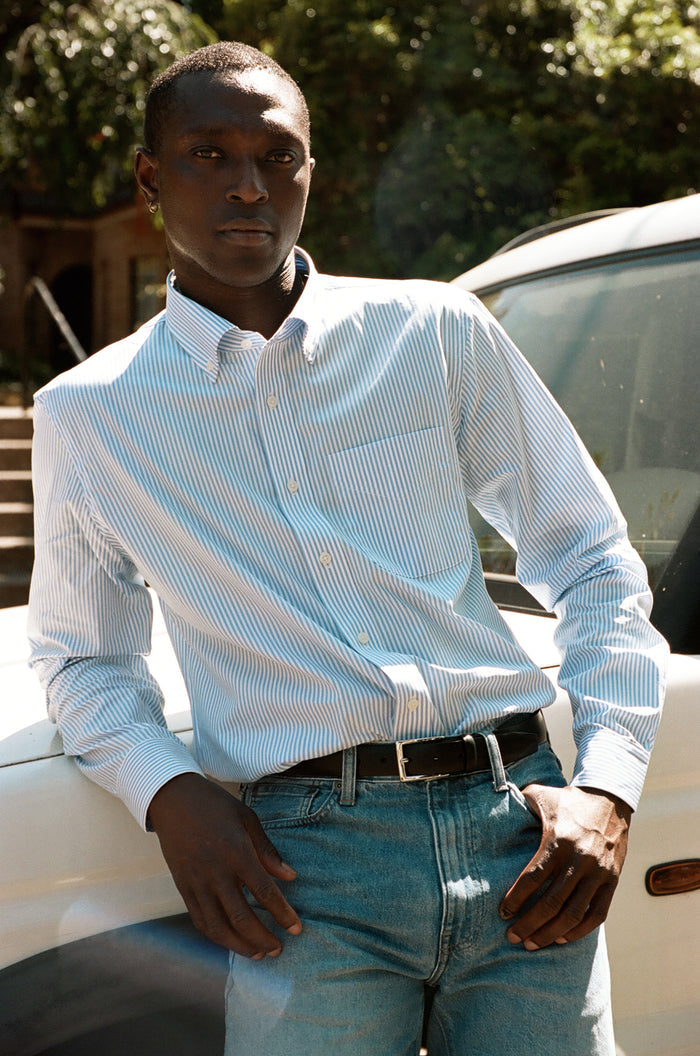 Man in a blue and white striped shirt and jeans leaning against a white SUV outdoors.