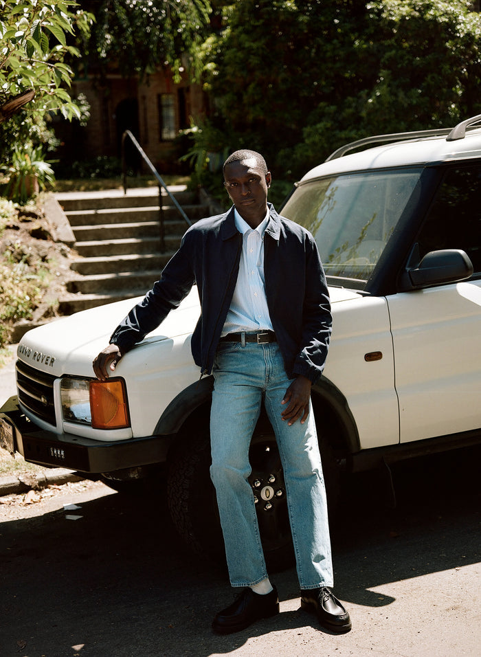 Man in a navy jacket over a blue and white striped shirt with jeans, beside a white Land Rover.