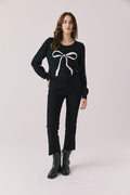 Full body view of woman wearing Chaser’s true black Bow Long Sleeve with white bow design, black jeans, and boots on a plain background.