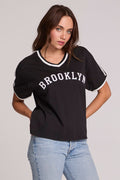 Front view of a woman wearing the Chaser Brooklyn Varsity Graphic Tee on a neutral background with hands on hips