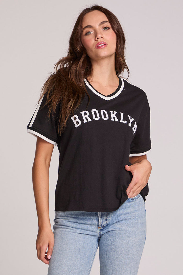 Front view of a woman wearing the Chaser Brooklyn Varsity Graphic Tee on a neutral background with one hand on hip