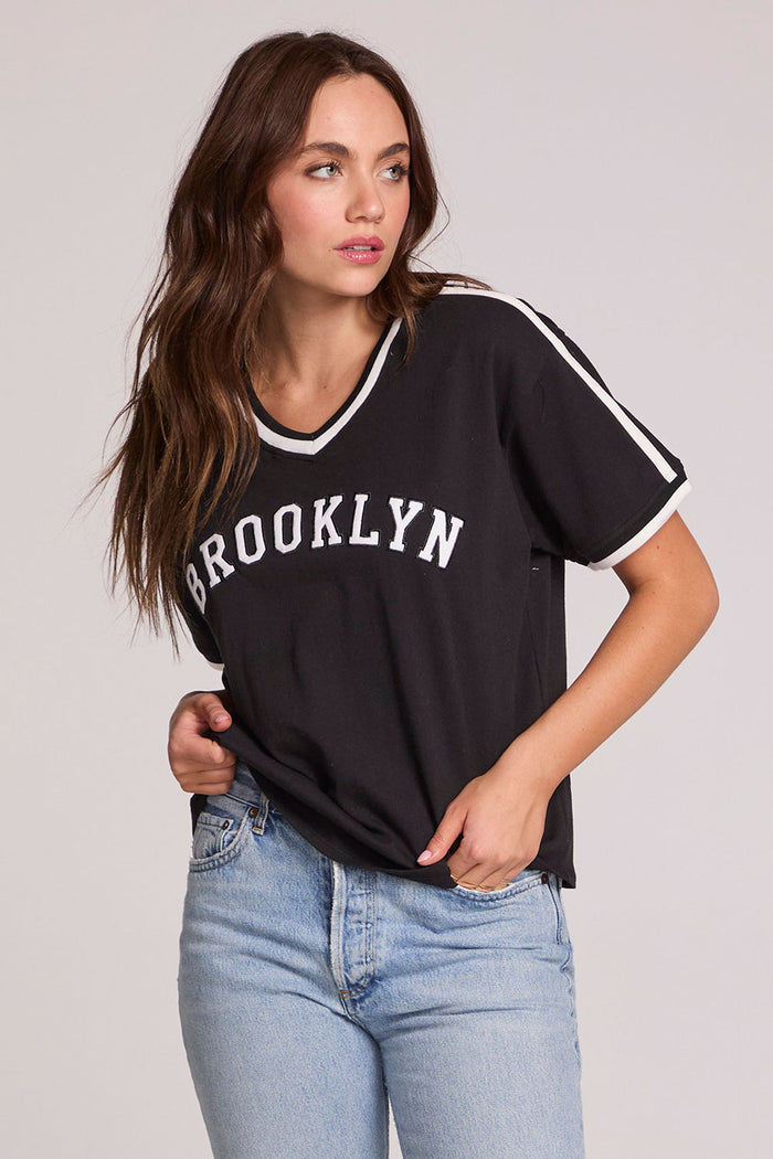 Front view of a woman wearing the Chaser Brooklyn Varsity Graphic Tee on a neutral background with one hand in her pocket