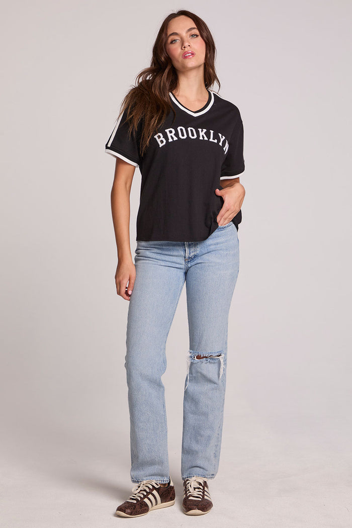 Full body front view of a woman wearing the Chaser Brooklyn Varsity Graphic Tee on a neutral background with one hand at her side