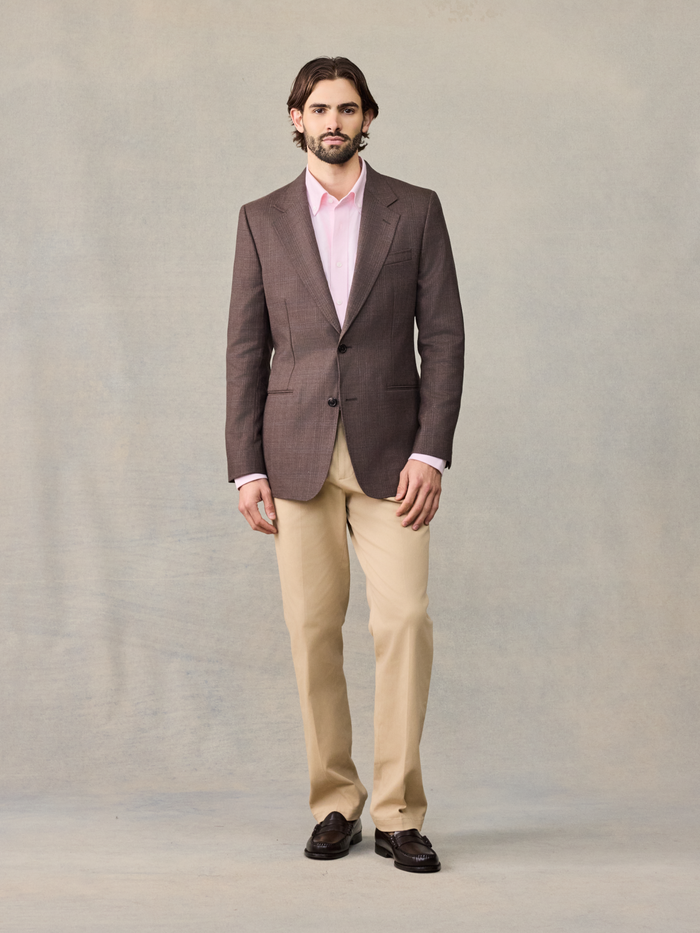 Man in a brown plaid sportcoat, pink shirt, and tan trousers, full body front.