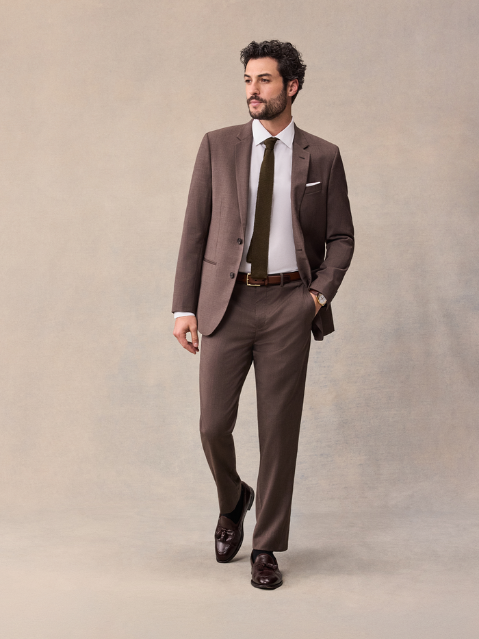 Man in an open brown suit jacket with white shirt and olive tie, full body front.