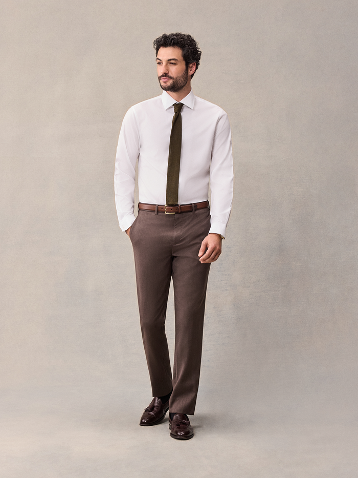 Man in a white shirt and olive tie wearing brown suit trousers, no jacket, full body.