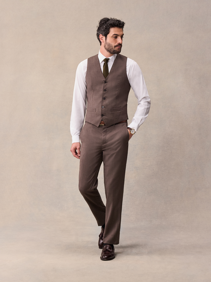 Man in a brown suit vest and trousers with a white shirt and olive tie, walking, full body.