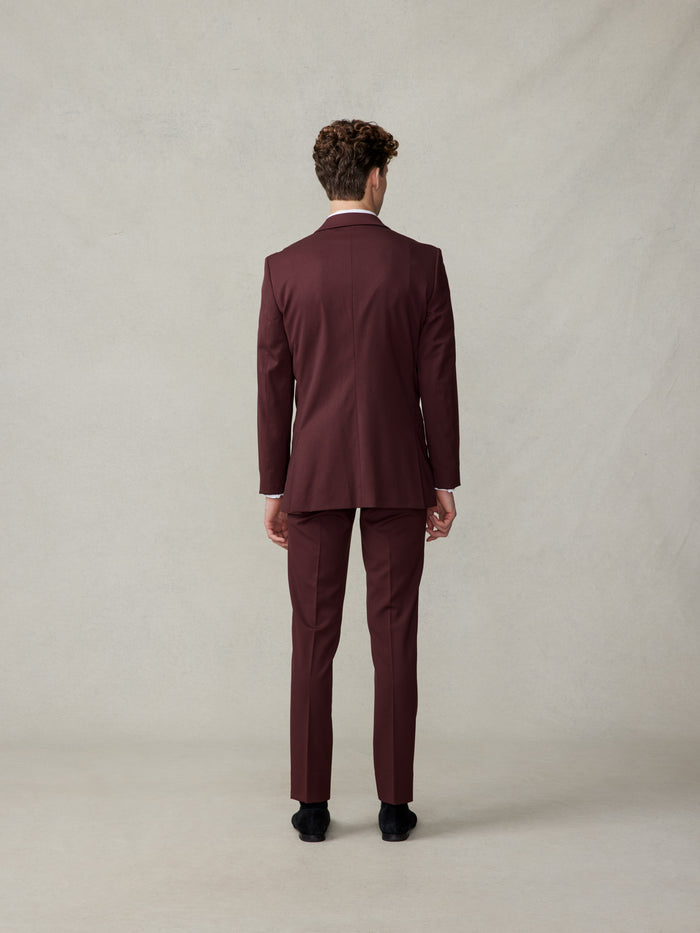 Man in a burgundy tuxedo viewed from the back, standing against a neutral background.