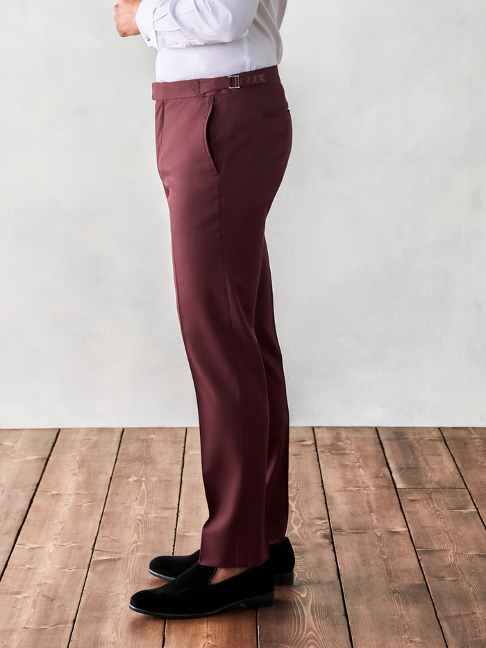 Side view of a man in burgundy tuxedo trousers with a side buckle strap and black velvet loafers.