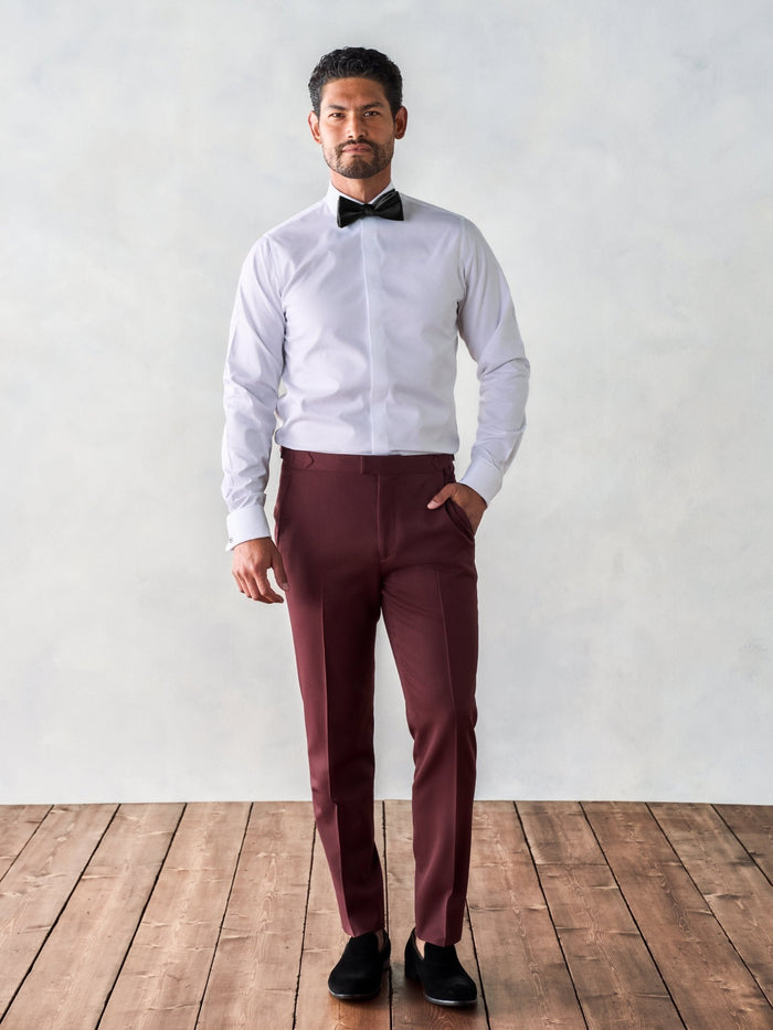 Man in a white tuxedo shirt and black bow tie wearing burgundy trousers, no jacket.