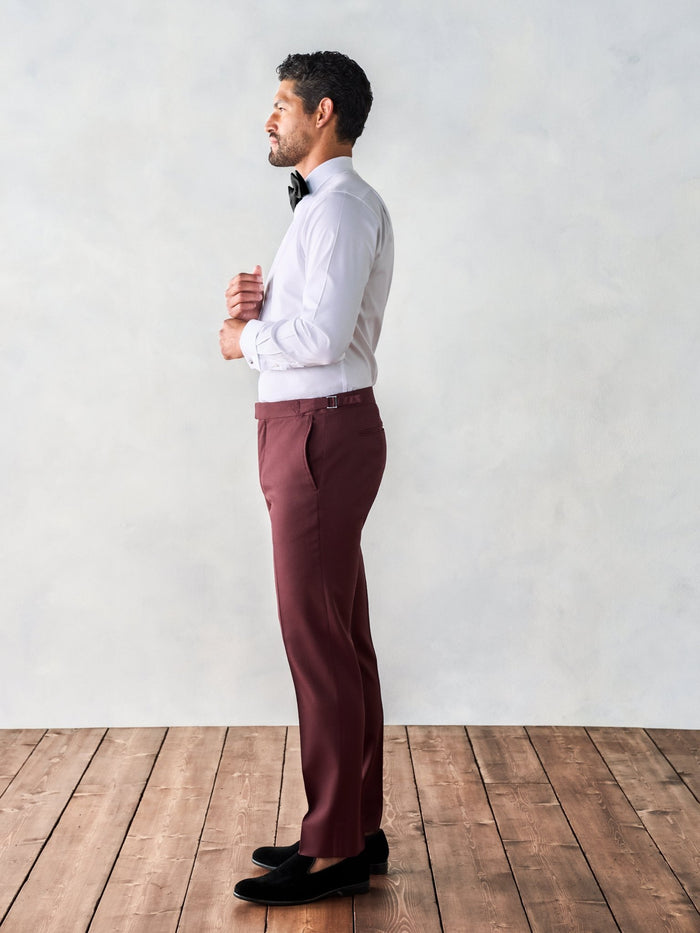 Side view of a man in a white tuxedo shirt and black bow tie wearing burgundy trousers.