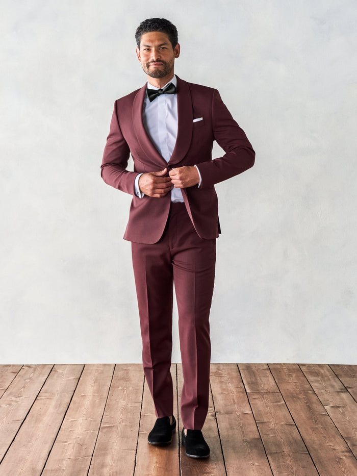 Man in a burgundy shawl-collar tuxedo buttoning the jacket, full body front, black bow tie.