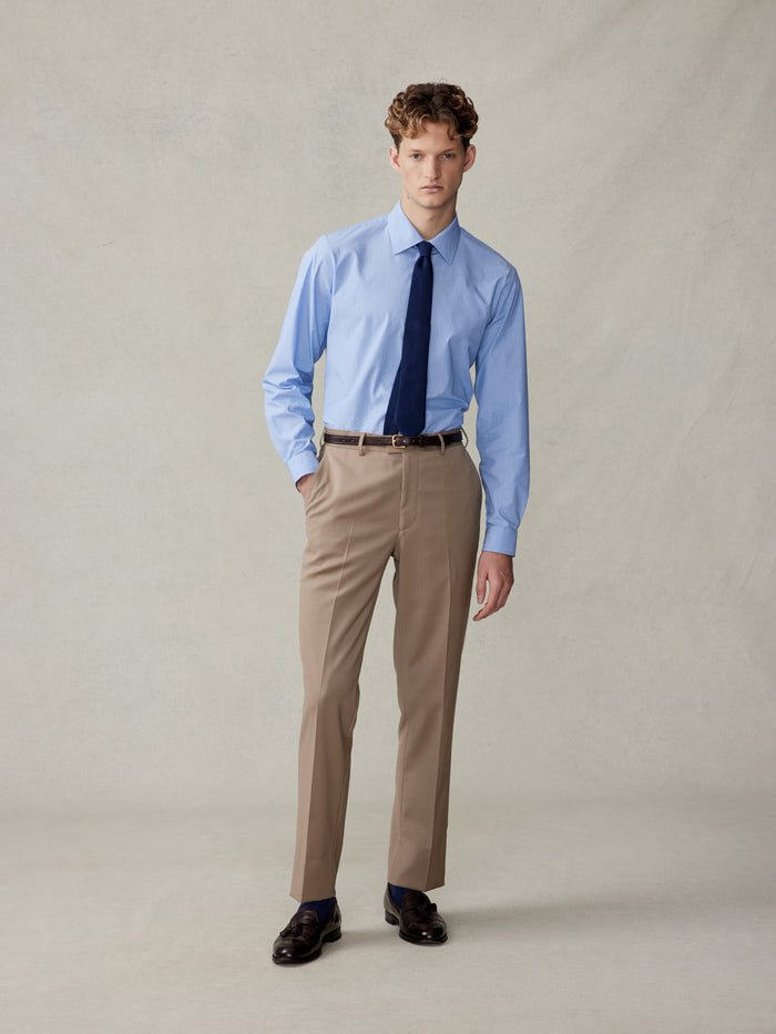 Man in a light blue shirt and navy tie wearing camel suit trousers, no jacket, full body front.