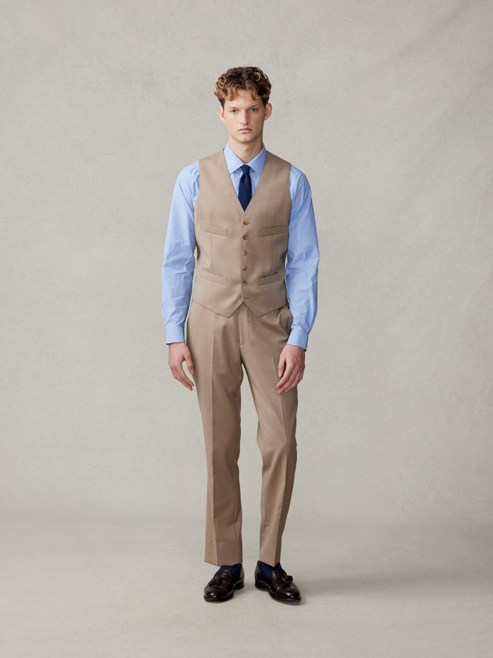 Man in a camel suit vest, light blue shirt, and navy tie, full body front.