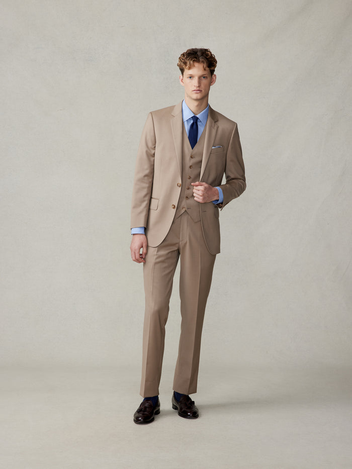 Man in a camel three-piece suit with a vest, light blue shirt, and navy tie, buttoning the jacket.