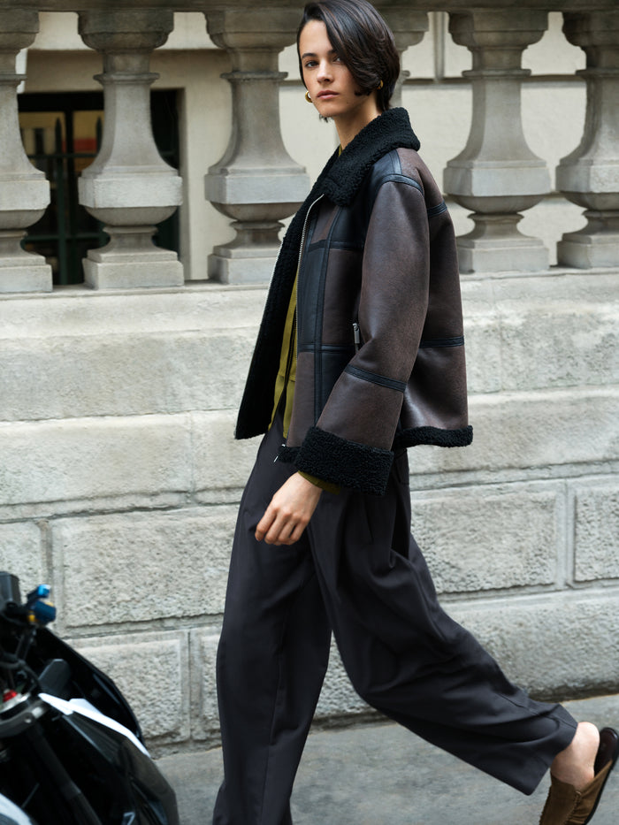 A woman wearing a brown faux suede aviator jacket with a faux shearling trim, zip through jacket, and side pockets.