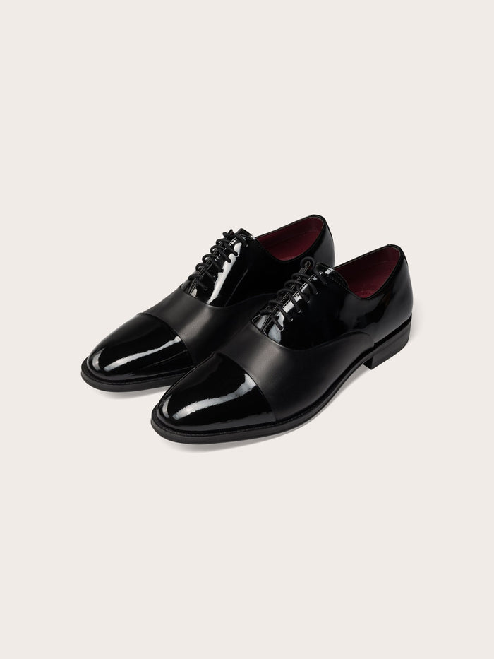 Pair of black cap-toe dress shoes in patent and calf leather, angled front view.