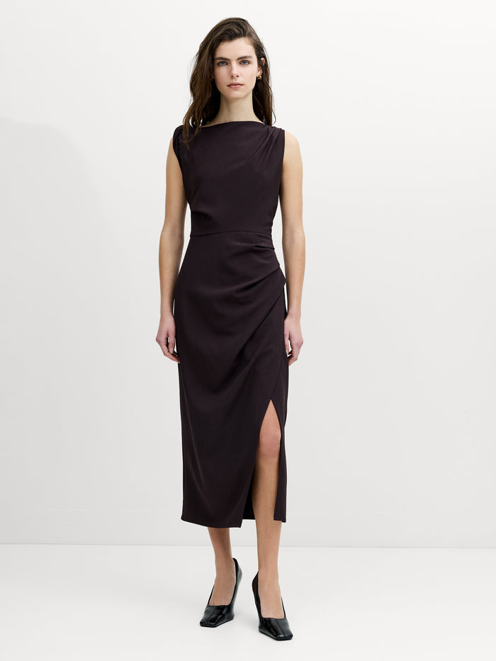A woman wearing a sleeveless, draped midi mocha brown dress with a straight neckline and a side slit hem.