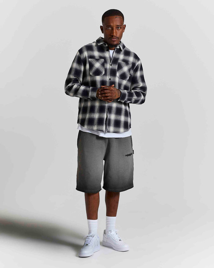 A man modeling a black and white plaid button-up shirt, dark gray shorts, white socks, and white sneakers against a light gray studio background.