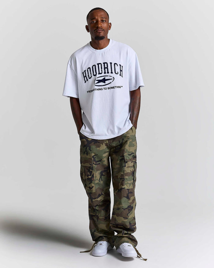 A man wearing a white HOODRICH t-shirt with a black graphic print, paired with green and brown camouflage cargo pants and white sneakers, standing against a light gray studio background.