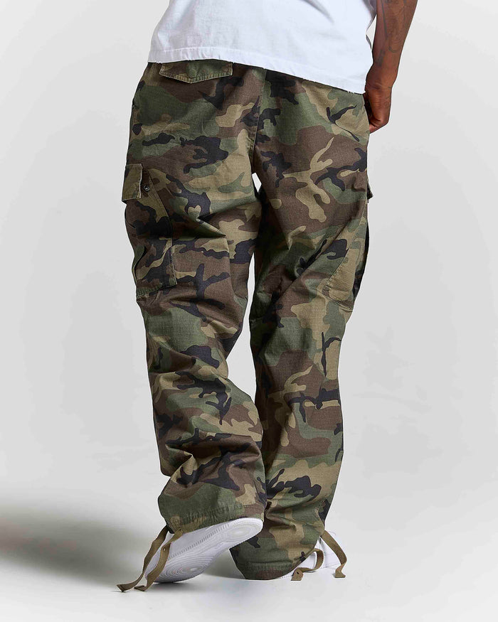 A person shown from the waist down wearing loose-fit camouflage cargo pants with a white t-shirt and white sneakers, photographed against a light gray studio background.