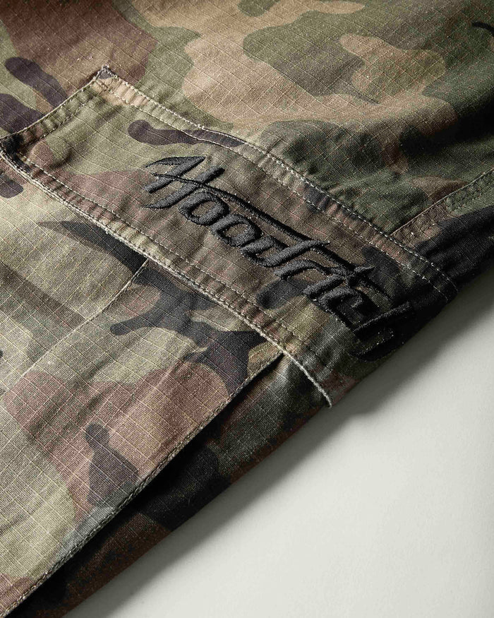 Close-up of a camouflage garment, likely a jacket or overshirt, showing a pocket with black embroidered script reading 'hoodRICH'. The fabric has a woodland camo pattern in green, brown, tan, and black.
