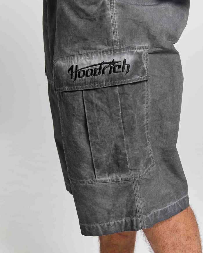 Close-up of a person wearing gray washed cargo shorts with a large front flap pocket and black embroidered 'Hoodrich' logo on the pocket, against a light gray background.