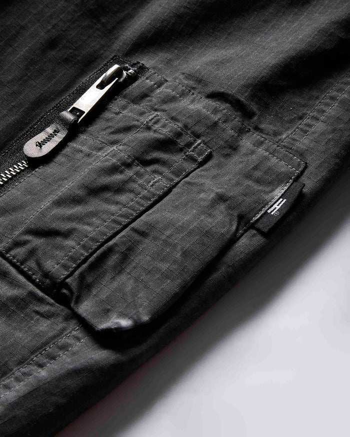 Close-up of a dark gray textured fabric garment showing a zippered pocket with a silver metal zipper pull and a small black brand tab.