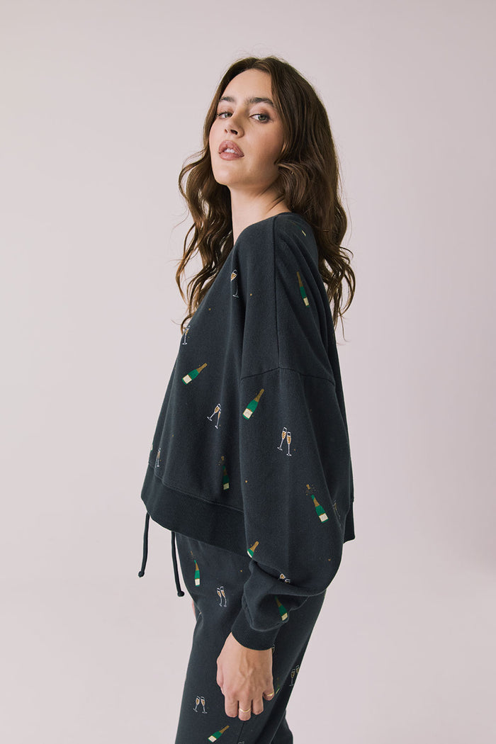 Side view of woman wearing Chaser’s black Champagne Party Long Sleeve Pullover with allover champagne bottle and glass graphics on a light grey background