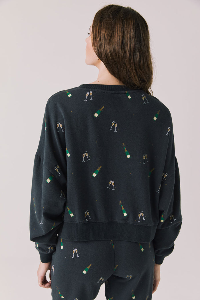 Back view of woman wearing Chaser’s black Champagne Party Long Sleeve Pullover with allover champagne bottle and glass graphics on a light grey background