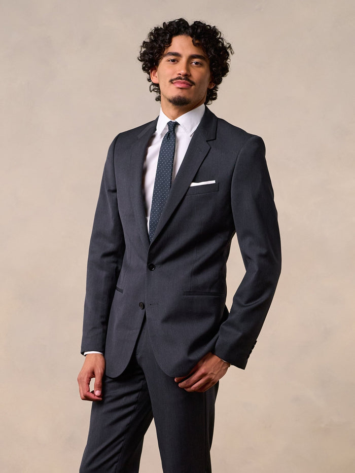 Man in a charcoal suit, white shirt, and dotted tie, front view cropped at the thighs.