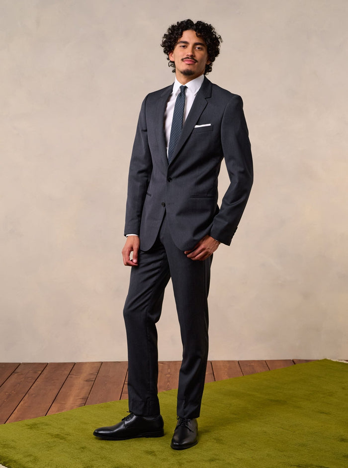Man in a charcoal suit with a white shirt and dotted tie, full body front, arms at sides.