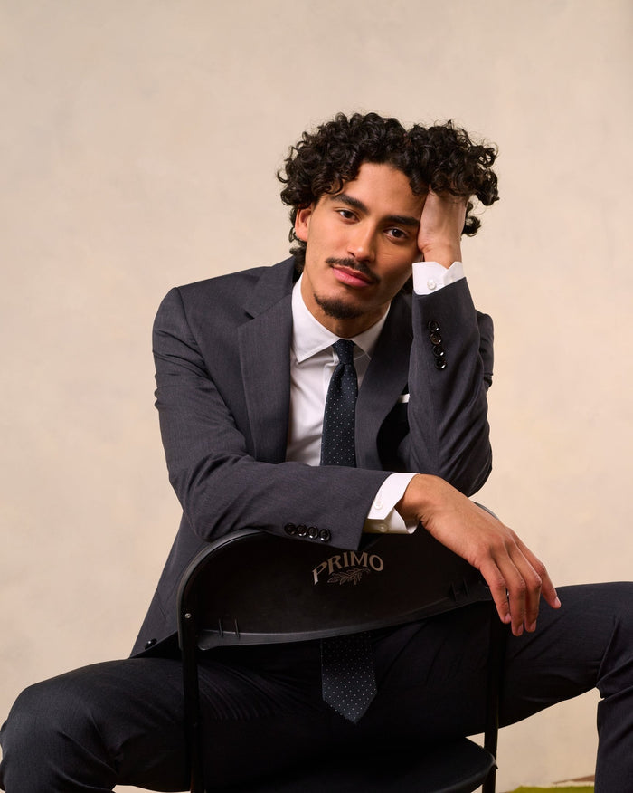 Man in a charcoal suit seated on a black folding chair, resting his head on his hand.