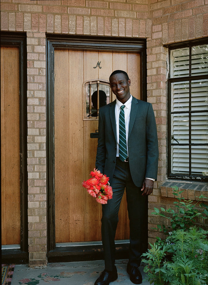Man in a charcoal suit holding a bouquet of red flowers, standing by a wooden door.