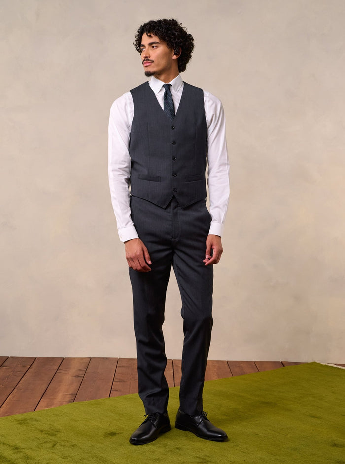 Man in a charcoal suit vest and trousers with a white shirt and tie, full body front.