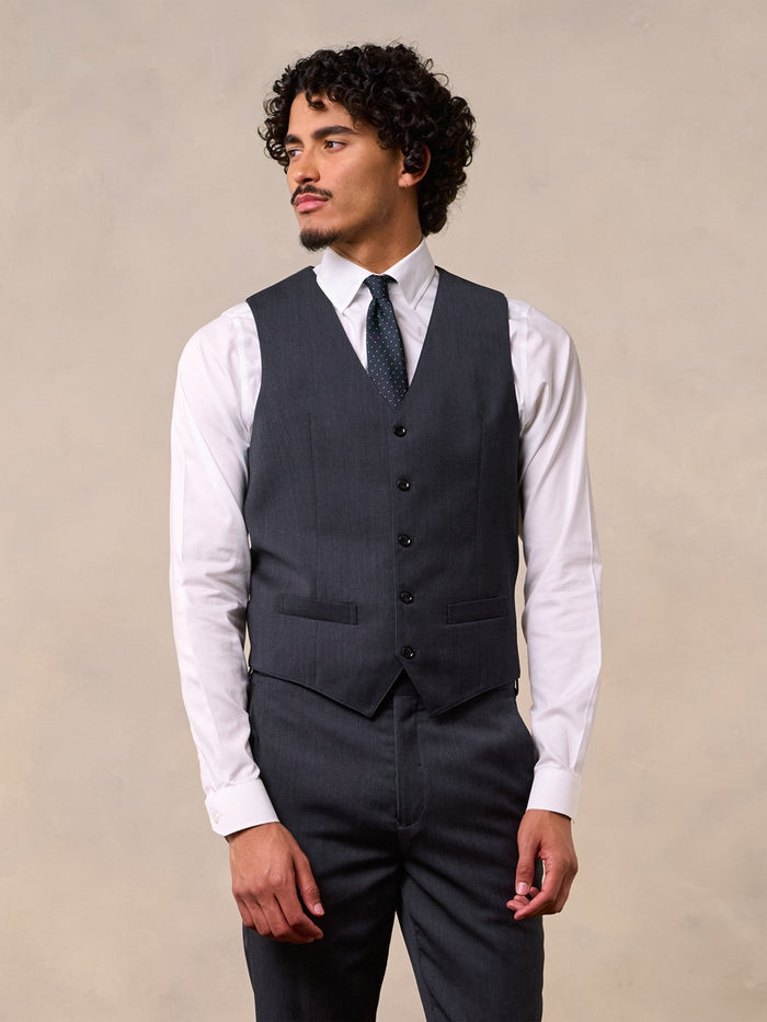A man in a charcoal suit vest over a white shirt with a navy dotted tie