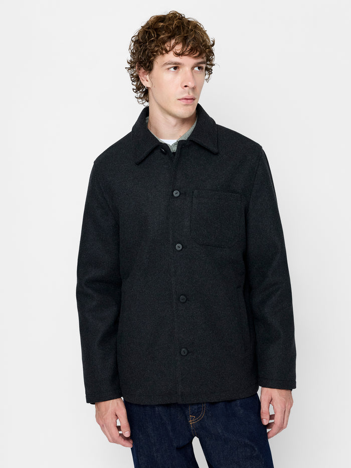 A man wearing a dark grey jacket with a single chest pocket, a button down fastening, long sleeves and a collared neckline.