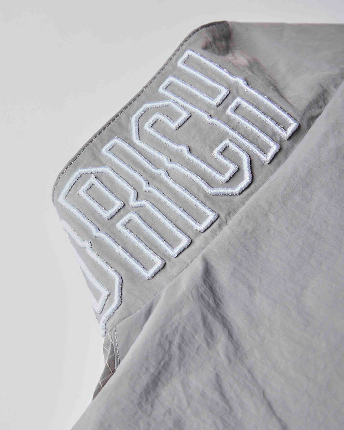Close-up of a light gray fabric garment with large white embroidered lettering reading 'RICH' on the shoulder.