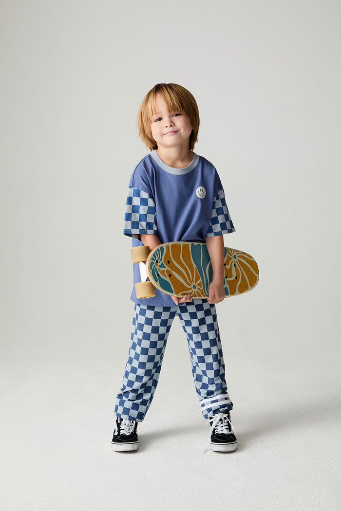 Front view of a boy wearing the Chaser Smileys Boys Tee on a neutral background with matching checkered pants holding long board while smiling