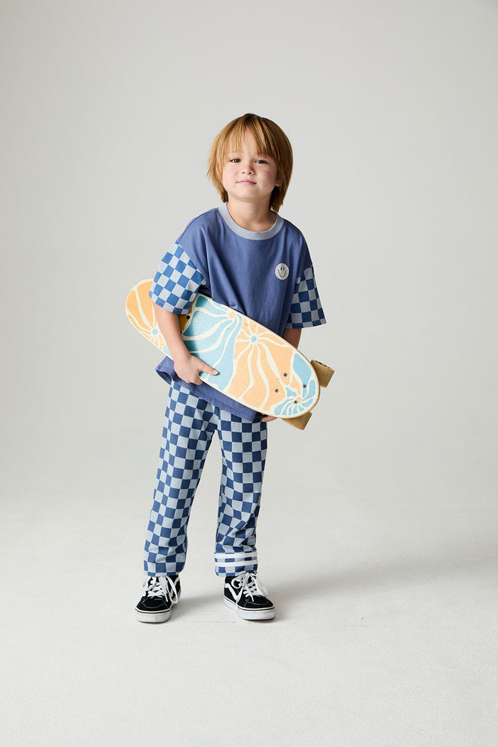 Front view of a boy wearing the Chaser Smileys Boys Tee on a neutral background with matching checkered pants holding long board