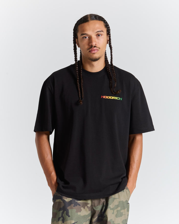 A man with long braids wearing a black HOODRICH t-shirt with a small multicolor logo on the chest, paired with camouflage pants, against a light gray studio background.