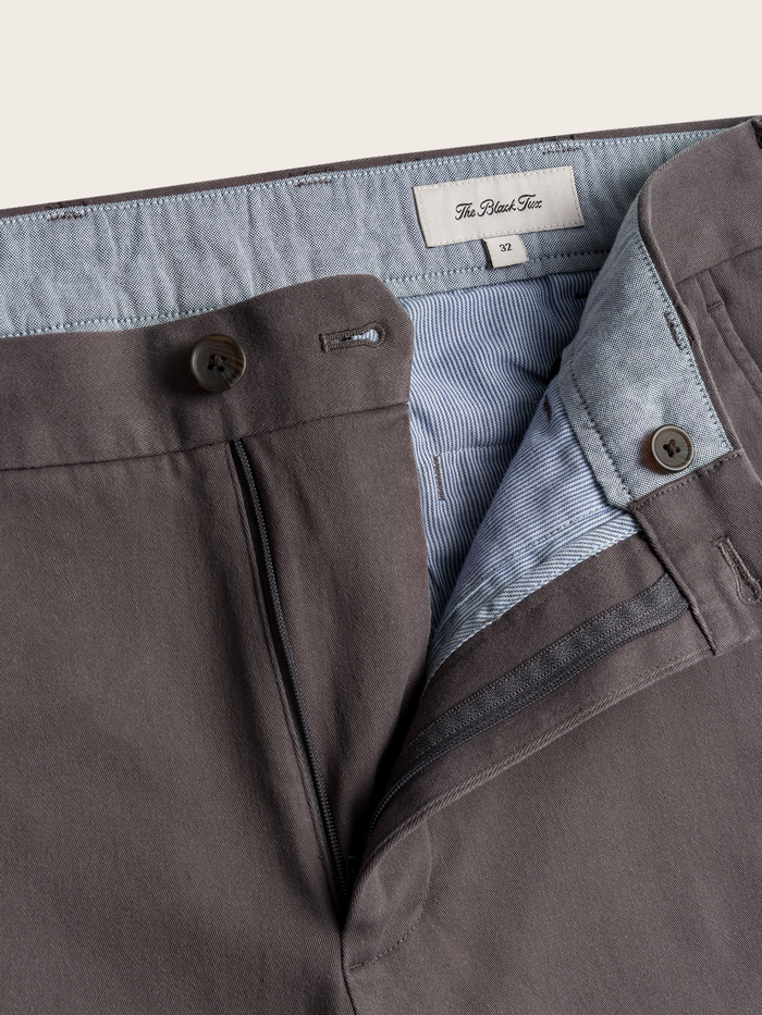 Close-up of grey chino waistband, unbuttoned fly, brand label and striped interior lining.