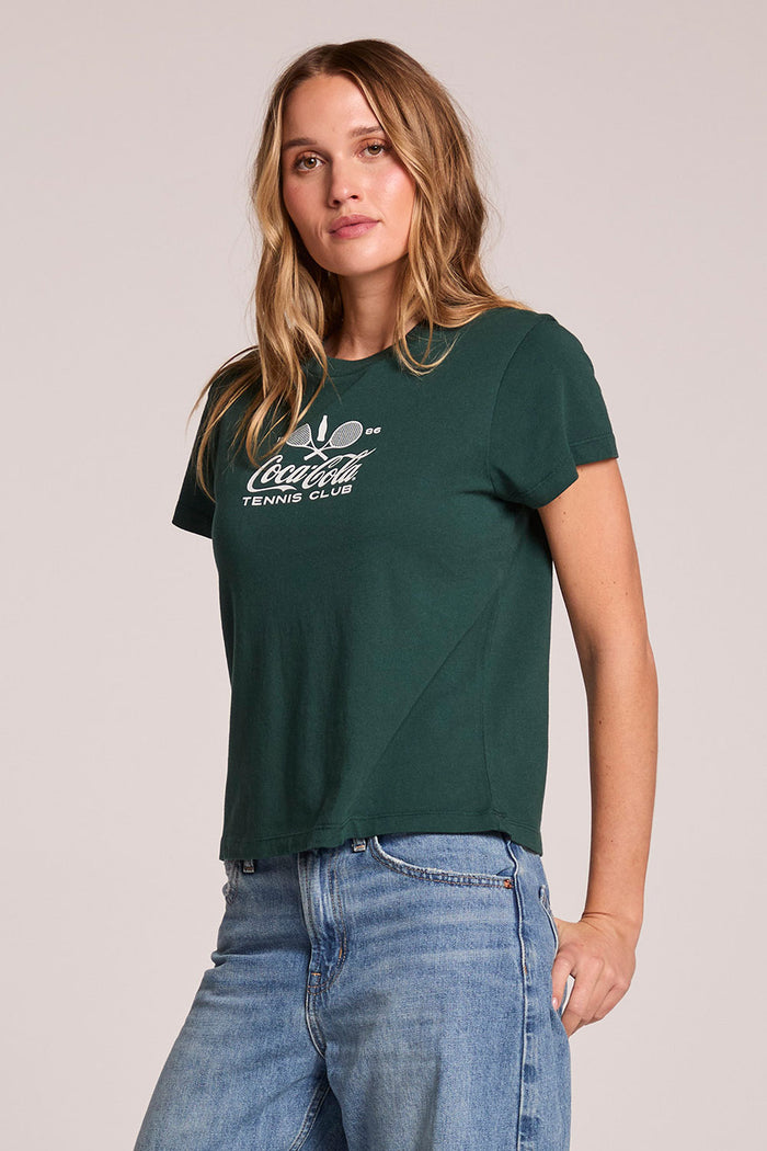 Side angle view of a woman wearing the Chaser   Coca Cola Tennis Club Tee on a neutral background with hands at her arms