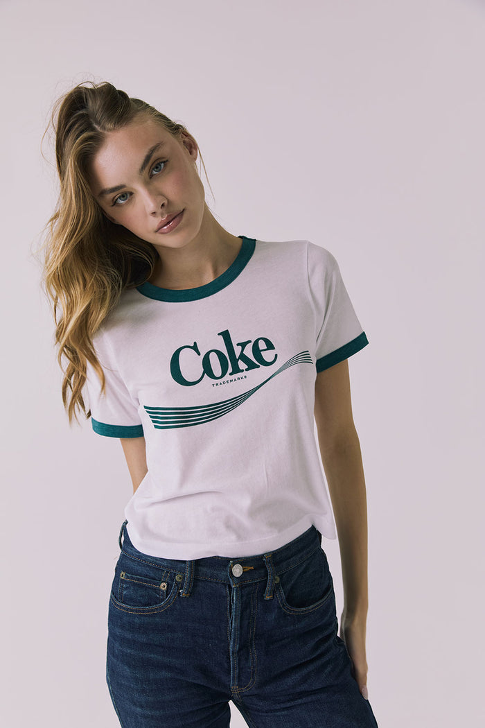Front view of a woman wearing Chaser's white Coca Cola The Real Thing tee with 'Coke' logo and blue jeans on a plain background