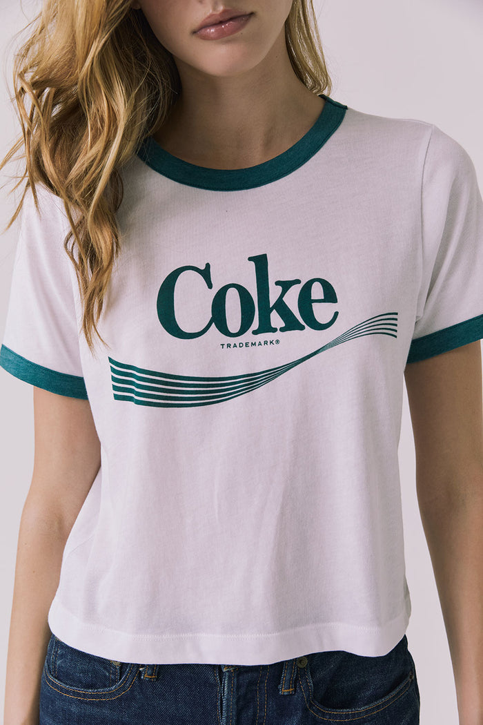 Close up front view of a woman wearing Chaser's white Coca Cola The Real Thing tee with 'Coke' logo and blue jeans on a plain background