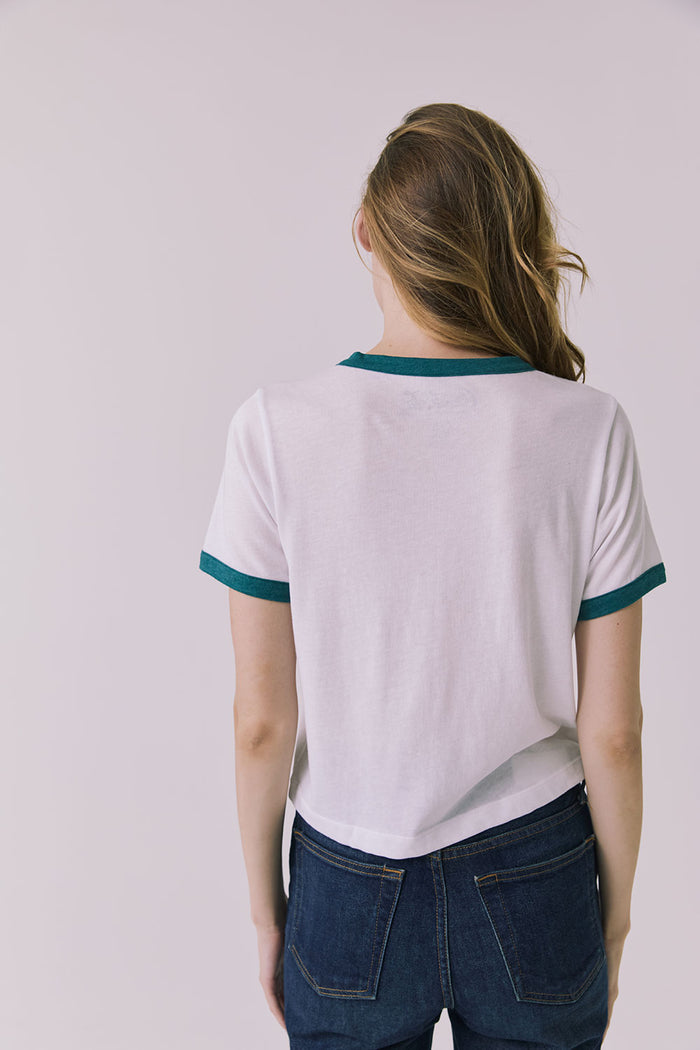 Back view of a woman wearing Chaser's white Coca Cola The Real Thing tee and blue jeans on a plain background