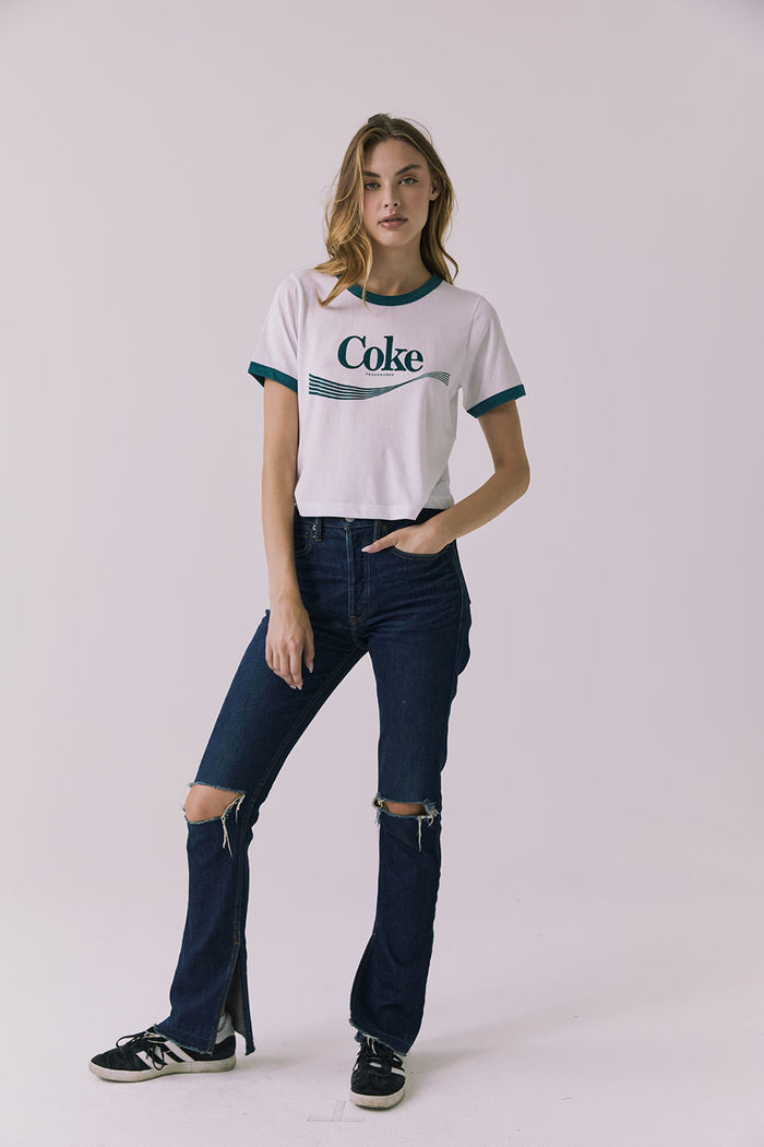 Full body front view of a woman wearing Chaser's white Coca Cola The Real Thing tee with 'Coke' logo and blue jeans on a plain background